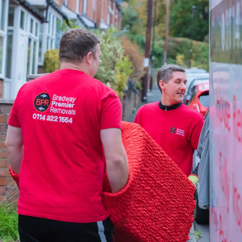 Two Bradway Premier Removals crew members carrying red wrapped furniture along a sloping terraced street to the van during a house removal