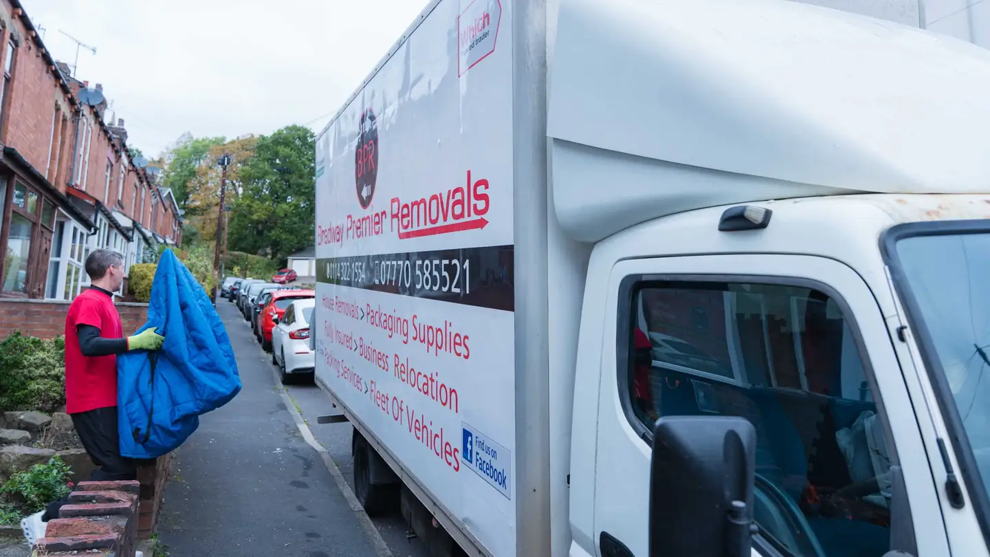 Bradway Premier Removals crew member carrying a blue furniture blanket beside the removals lorry on a sloping terraced street