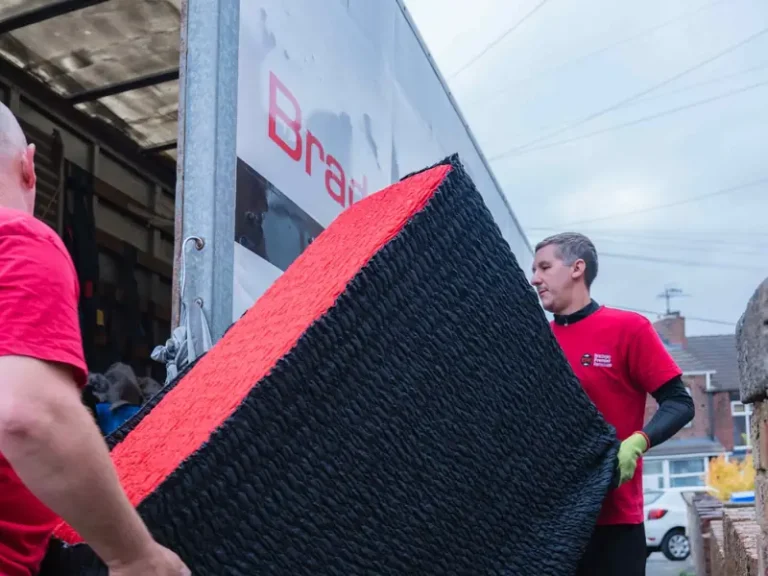 Two Bradway Premier Removals crew members carrying black and red wrapped furniture beside the van on a terraced street