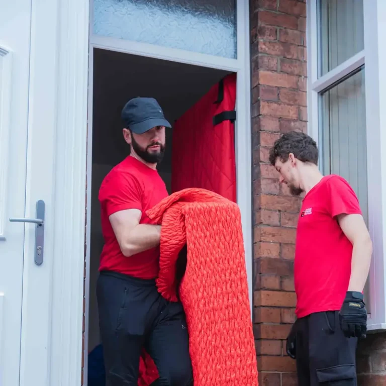 Two Bradway Premier Removals crew members carrying red quilted wrapped furniture out of the front door of a brick terraced house