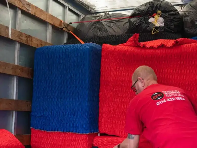 Bradway Premier Removals crew member loading stacked red and blue wrapped furniture into the van